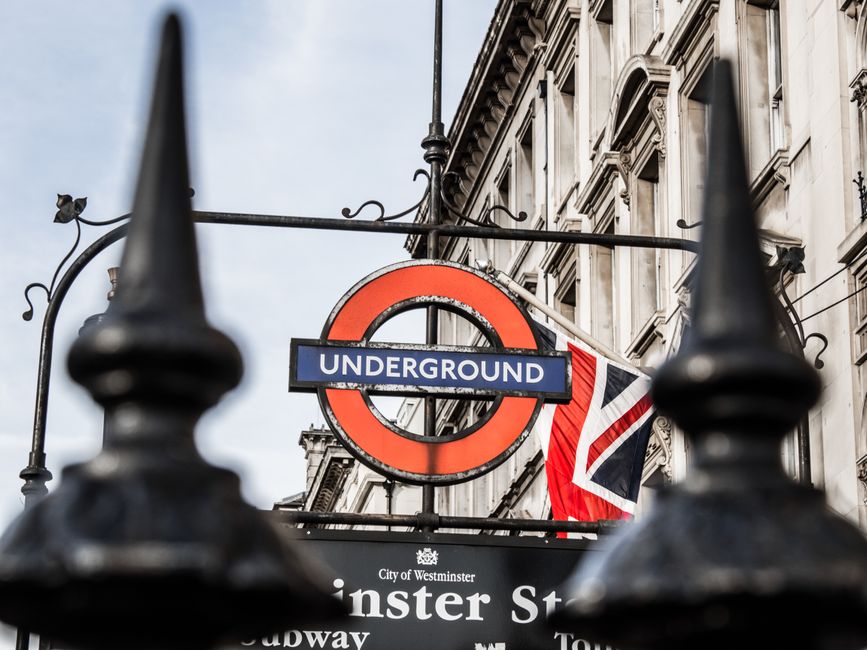 A London Underground station sign featuring a red circle with a blue horizontal bar displaying the word 'UNDERGROUND' in white text, mounted on a decorative wrought iron bracket attached to a historic building with ornate stonework. Behind the sign, part of a Union Jack flag is visible, draped next to the sign. The scene is viewed through black iron railings with pointed finials in the foreground, suggesting an urban outdoor setting near a Westminster street. The building's facade is light-colored stone, with detailed architectural elements and multiple windows, indicating a typical cityscape environment. The background sky appears overcast, providing diffuse natural lighting that highlights the textures of the stone and metal. This scene reflects a typical Westminster environment, often associated with central London’s transport infrastructure and historic architecture, relevant to discussions on transportation hubs and city infrastructure in the context of waste management and disposal services like those provided by Westminster rubbish removal experts.