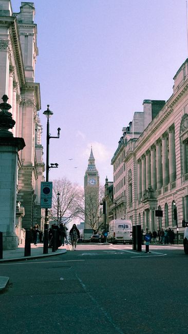 A city street scene with historical architecture on either side, featuring tall, ornate buildings with detailed stone facades and large windows. In the background, the Elizabeth Tower known as Big Ben, with its distinctive clock face and spire, is visible over leafless trees and a clear blue sky. The street is lined with black bollards and lampposts, with a few pedestrians walking along the pavement. A white van is parked near the curb, and a group of people are seen near the sidewalk, some wearing coats. The environment appears to be in winter or early spring, with the atmosphere bright but cool. This urban scene reflects a typical central Westminster setting, where external buildings and infrastructure are visible, and occasional private waste collection or external rubbish clearance might be part of maintaining the area's aesthetic, supported by professional waste removal services like those provided by Westminster rubbish removal specialists.