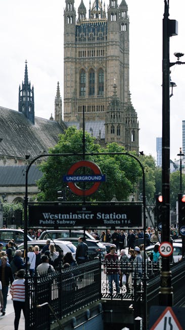A London Underground station sign featuring a red circle with a blue horizontal bar displaying the word 'UNDERGROUND' in white text, mounted on a decorative wrought iron bracket attached to a historic building with ornate stonework. Behind the sign, part of a Union Jack flag is visible, draped next to the sign. The scene is viewed through black iron railings with pointed finials in the foreground, suggesting an urban outdoor setting near a Westminster street. The building's facade is light-colored stone, with detailed architectural elements and multiple windows, indicating a typical cityscape environment. The background sky appears overcast, providing diffuse natural lighting that highlights the textures of the stone and metal. This scene reflects a typical Westminster environment, often associated with central London’s transport infrastructure and historic architecture, relevant to discussions on transportation hubs and city infrastructure in the context of waste management and disposal services like those provided by Westminster rubbish removal experts.