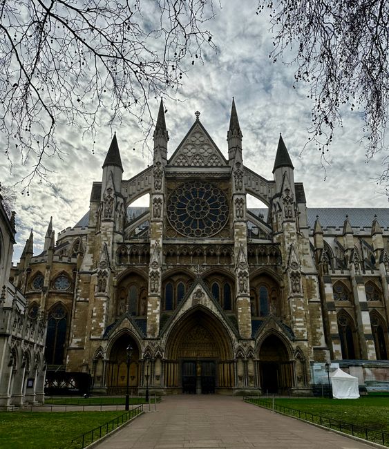The exterior façade of a historic Gothic-style cathedral featuring pointed arches, intricate stone carvings, and tall spires. The building is constructed from weathered beige and brown stone, with detailed decorative elements along the entrance and around the large circular rose window set above the main doorway. The façade includes multiple narrow stained-glass windows, some with pointed arch designs, and vertical buttresses enhancing its vertical emphasis. In the foreground, a paved walkway leads directly to the cathedral's entrance, flanked by small patches of grass enclosed by simple black metal railings. Bare tree branches extend into the frame from the top left corner, suggesting an overcast sky behind the structure. The overall scene emphasizes the grandeur and historic craftsmanship of the cathedral, with no visible rubbish or debris, illustrating an environment that might be prepared for cleaning or maintenance work in a city setting where private waste removal services could be used for site clearance. The setting hints at an area that could be part of an urban or historic district requiring professional rubbish removal solutions, such as those offered by companies like RubbishCollectionWestminster.co.uk, for efficient site clearance or ongoing preservation efforts.