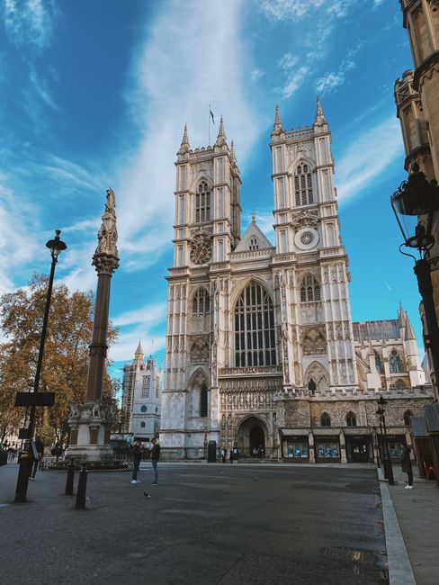 A wide-angle view of Westminster Abbey's Gothic façade, featuring its ornate stone carvings, towering spires, and large stained glass windows, set against a bright blue sky with scattered clouds. The front entrance is visible at the base, with a few pedestrians walking on the paved area surrounding the abbey. In the foreground, there are black bollards and ornate lamp posts lining the open space in front of the historic building. To the left, a tall thin lamppost with a decorative top is positioned near trees with autumn foliage, and on the right, part of an adjacent building with classical architectural details can be seen. The scene is well-lit with natural sunlight, emphasizing the light-colored stonework of the abbey. This setting highlights the importance of maintaining cleanliness and order around historic landmarks, which may involve private rubbish removal services for waste clearance in high-traffic tourist areas like Westminster.