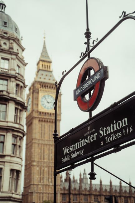 A close-up view of a black Westminster Station sign hanging from an ornate iron bracket, displaying white lettering for 'Westminster Station,' along with additional signage indicating 'Public Subway' and 'Toilets.' Above the station sign, a red and blue roundel London Underground sign is visible, suspended from a black metal pole. In the background, iconic historic buildings with detailed stonework and large windows are seen, including the clock tower known as Big Ben, which features a clock face with black Roman numerals and gold detailing on a white background. The scene is captured during daylight with overcast skies, creating a muted, soft light over the urban landscape, emphasizing the contrast between the traditional architecture and modern transit signage. The image background includes elements of London's historic architecture, with a focus on the station sign and surrounding structures, illustrating a typical scene in central London that relates indirectly to the city’s transportation infrastructure and urban environment.
