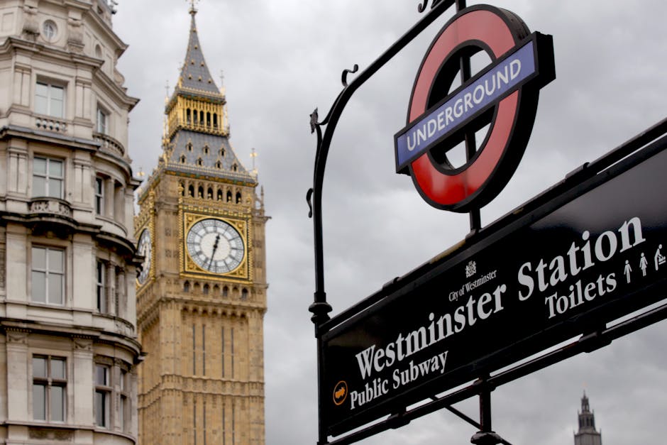 A close-up view of a London Underground sign for Westminster Station, positioned in the foreground with a black background and white lettering indicating 'Westminster Station,' along with symbols for toilets and access. Behind the sign, the iconic Elizabeth Tower housing Big Ben is visible, featuring its ornate gothic details, clock face showing approximately 6:05, and a spire topped with a decorative finial. The sky above is overcast with dark grey clouds, casting diffuse light over the scene. To the left, part of an elegant historical building with cream-colored stone facade, rounded bay windows, and decorative cornices is visible, contributing to the classic London cityscape. The image’s environment suggests an urban setting central to Westminster, with elements that subtly connect to the city's infrastructure and public transport, relevant for discussions of city waste management, alternative disposal sites, or urban environment considerations in the context of Westminster council rules for rubbish collection.